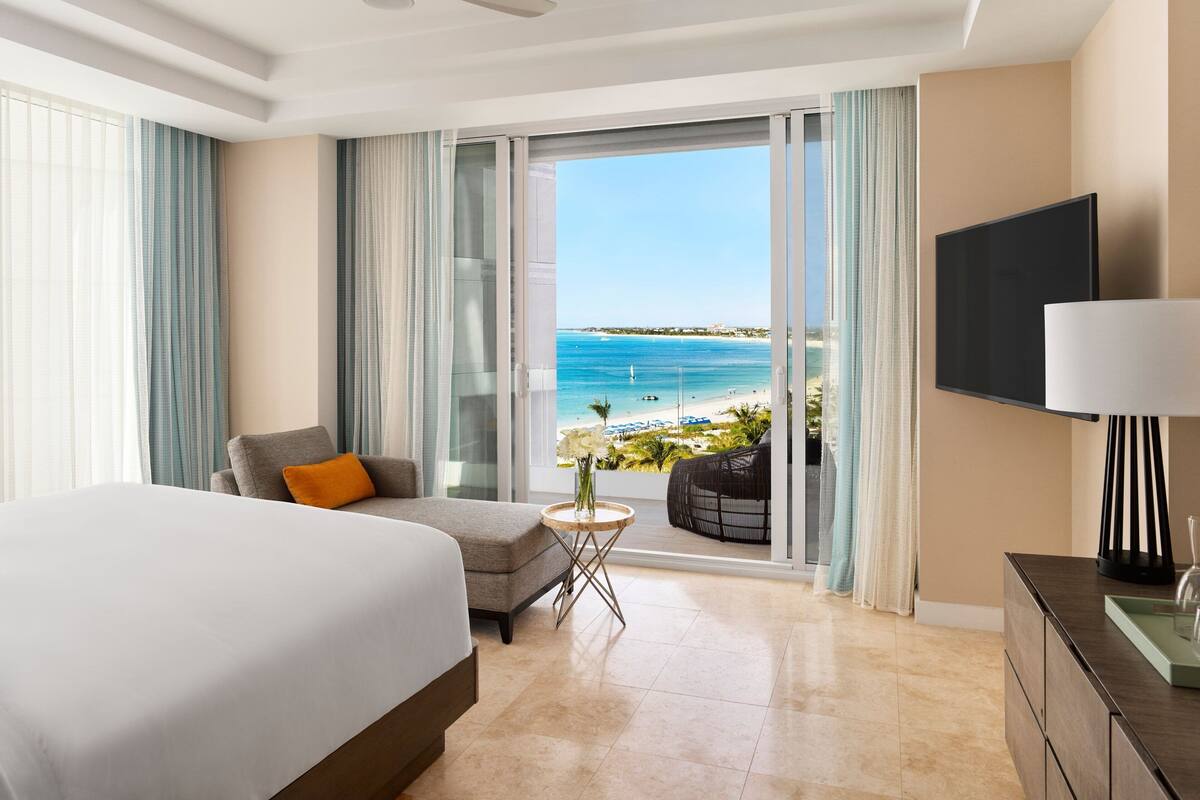 Ein Hotelzimmer mit Bett, einer Sitzecke, einem kleinen Tisch mit einer Vase, einem Fernseher und einem Balkon mit Blick auf Strand und Meer.