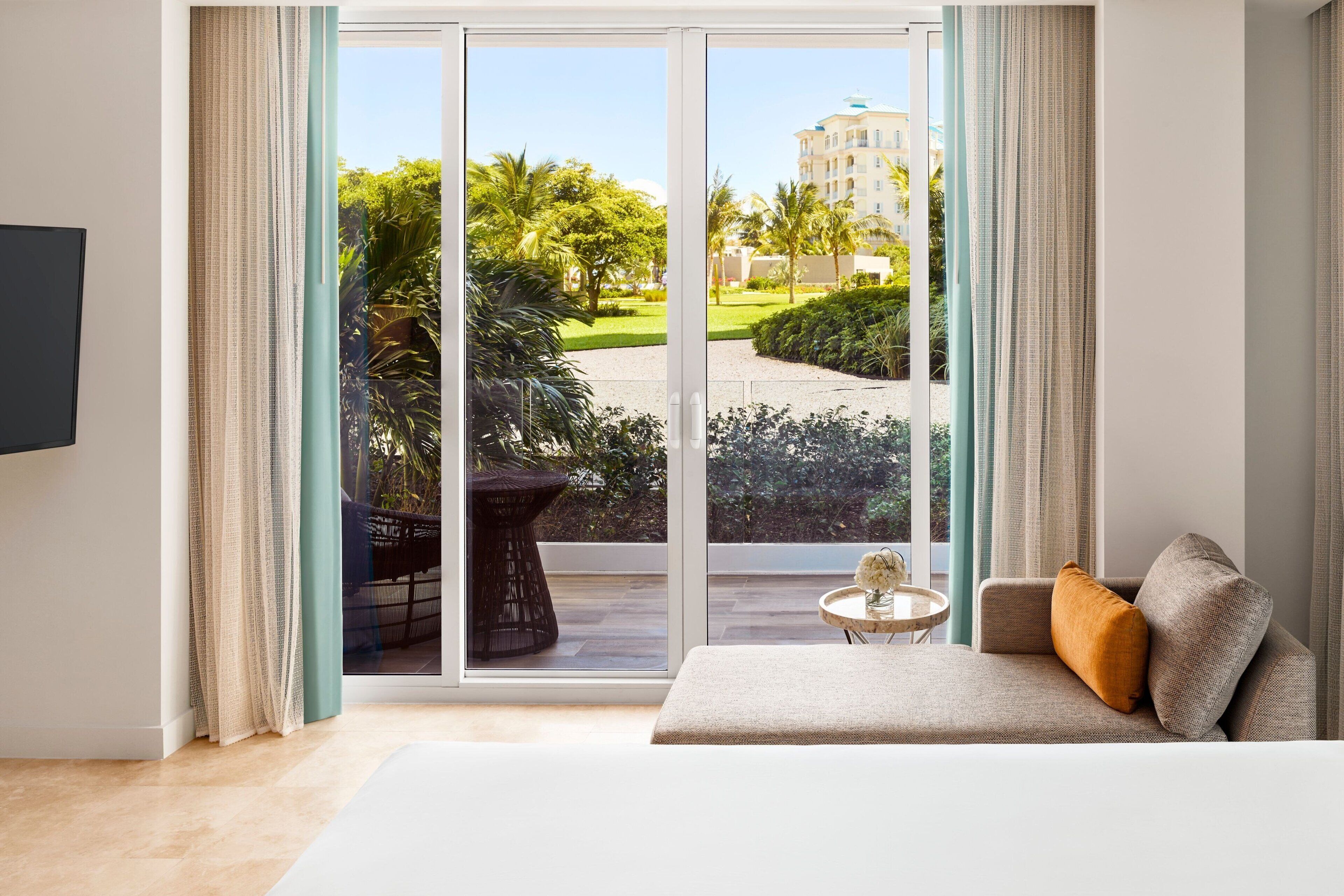 A modern living room with a sofa, a small round table, and a large sliding glass door leading to a balcony with a view of greenery and a building.