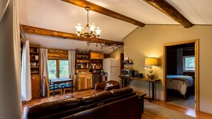 A cozy living room with a sofa, wooden beams, a chandelier, and a view of the kitchen and bedroom.