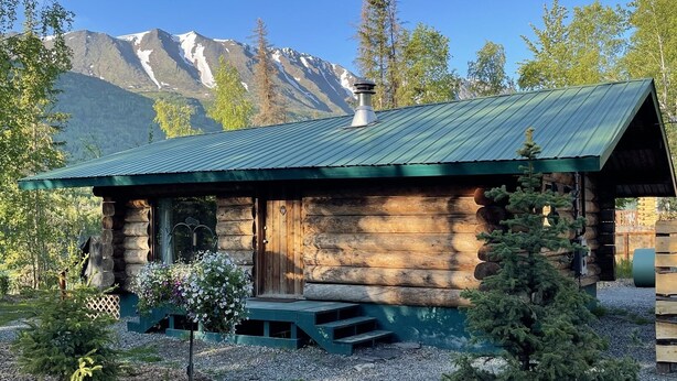 A rustic log cabin with a green metal roof, surrounded by a well-maintained garden with a small tree and a bench.
