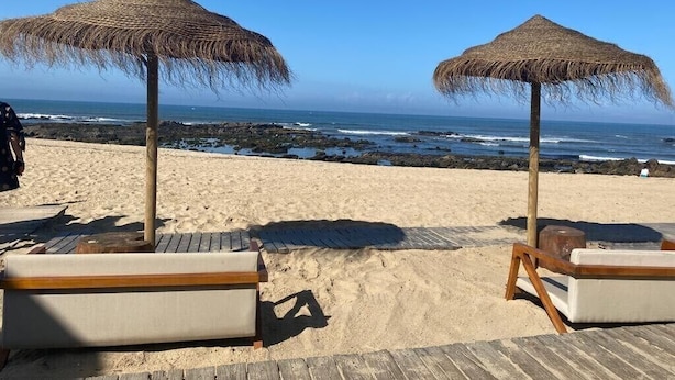 Two thatched umbrellas on a wooden deck overlooking a beach with rocky shoreline and ocean waves.