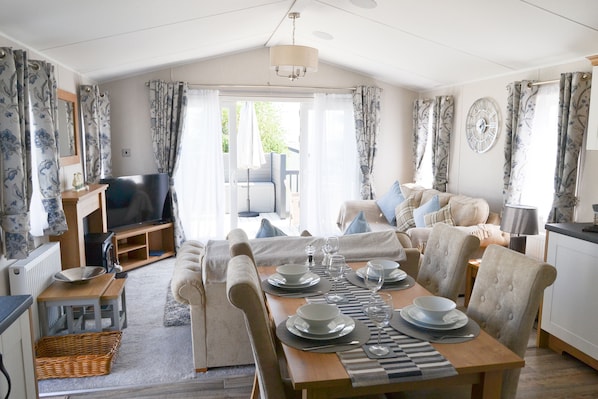 A well-lit living room with a sofa, dining table set for four, a television, and a large window with curtains.