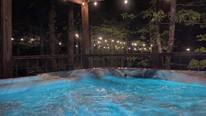 A hot tub with a wooden deck, surrounded by trees and string lights.