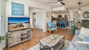 A modern living room with a TV, wooden entertainment unit, and a dining area with green chairs. - Maui Banyan G410 - Remodeled, Partial Ocean View, AC, Across Kamaole Beach! (Kihei)