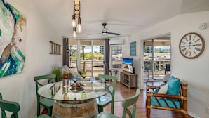 A modern living room with a glass dining table, green chairs, a wooden barrel, and a view of the outdoors. - Maui Banyan G410 - Remodeled, Partial Ocean View, AC, Across Kamaole Beach! (Kihei)