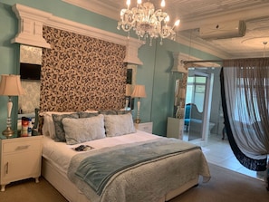 A bedroom with a bed, nightstands, a chandelier, a mirror, and a bathroom visible through a glass door.
