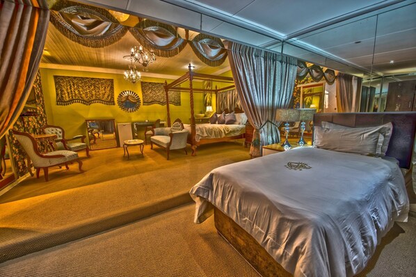 A spacious bedroom with a four-poster bed, a sitting area with chairs, and a large mirror reflecting the room.