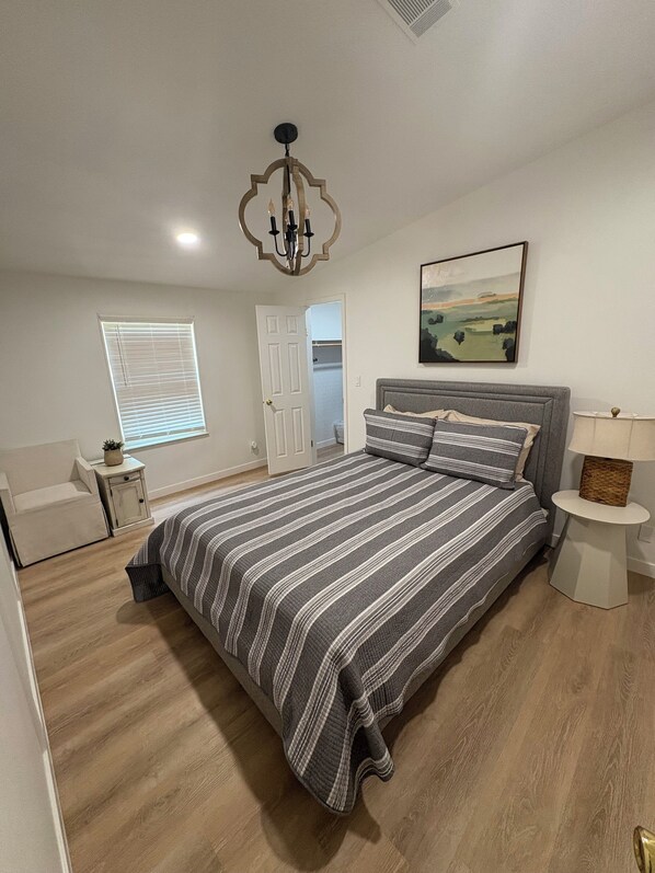 A bedroom with a large bed, a bedside table, a nightstand, a window with blinds, a door, a chandelier, and a painting on the wall. - Lake Fork Water Front, Privately Owned Dock - Lake levels are seasonally low. (Quitman)