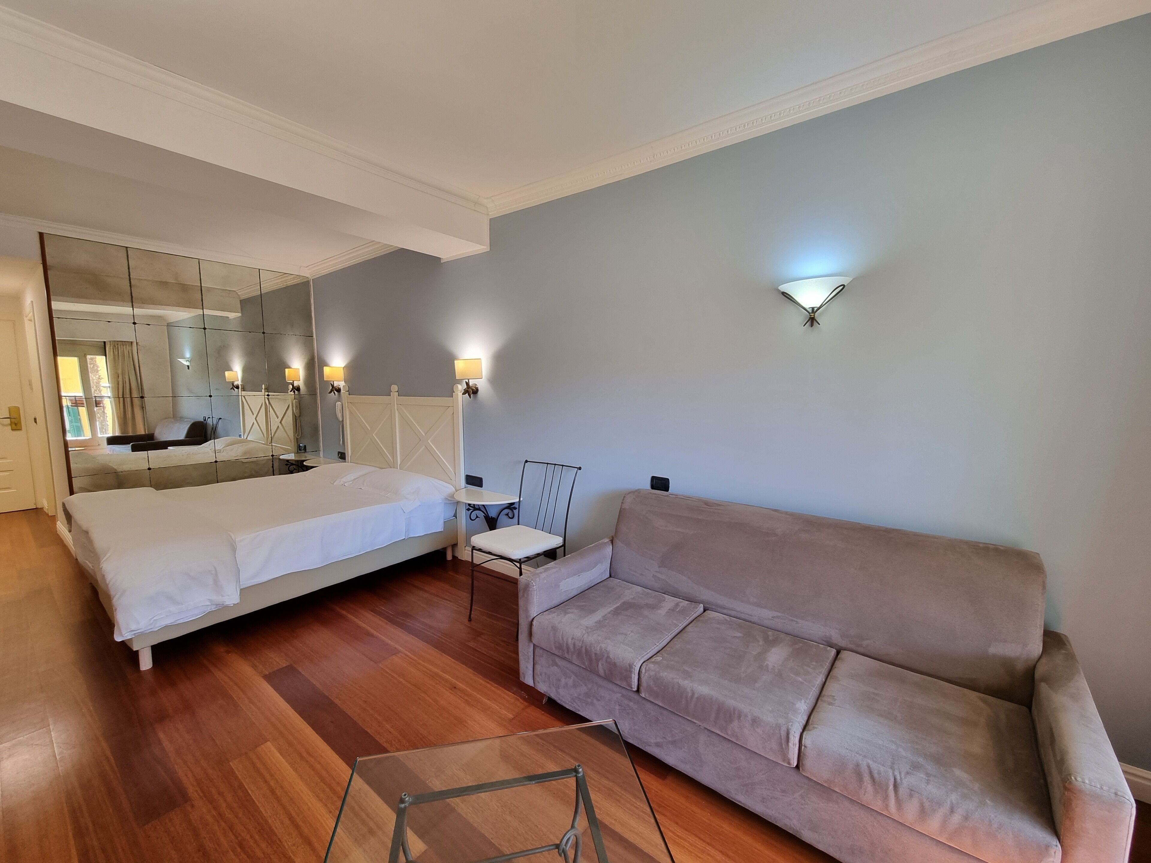 A hotel room with a large bed, a sofa, a small table, and a mirror.