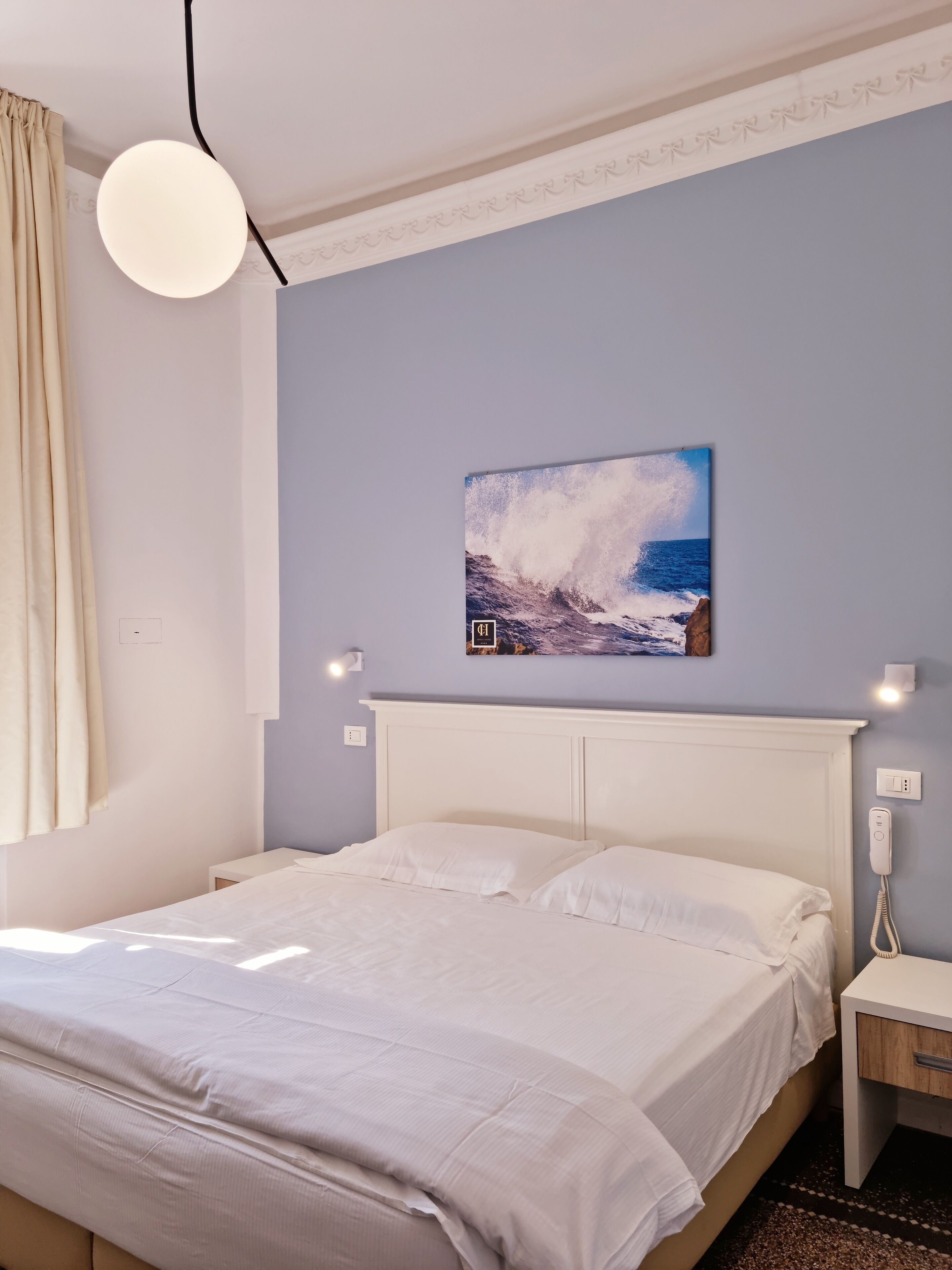 A bedroom with a bed, a nightstand, a wall-mounted phone, and a painting of a wave crashing against rocks.
