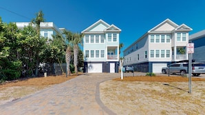 A two-story residential building with a garage, a paved driveway, and a fenced area with palm trees. - Paradise awaits! 7 Bedroom, Private Neighborhood Pool, Walk to the Beach! (Isle Of Palms)