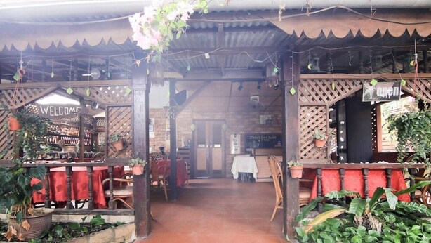 Um restaurante com um letreiro de 'WELCOME', estruturas de madeira treliçada e mesas ao ar livre com toalhas vermelhas.