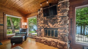 L’intérieur chaleureux d’un chalet avec une cheminée en pierre, un plafond en bois et de grandes fenêtres offrant une vue sur l’extérieur.
