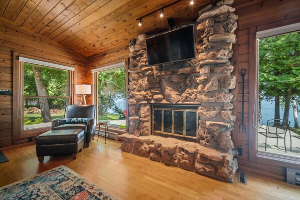 Interior acogedor de una cabaña con chimenea de piedra, techo de madera y ventanas amplias con vistas al exterior.