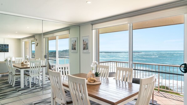 Un espace repas moderne avec une table en bois et des chaises blanches, de grandes fenêtres offrant une vue sur l’océan et un balcon doté d’une balustrade.