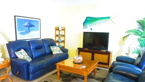 A living room with a blue sofa, a wooden coffee table, a flat-screen TV, and a decorative fish on the wall. - SUBTROPICAL OASIS CONDO, HEATED POOL, STEPS TO THE OCEAN ACCESS, GREAT LOCATION (Cocoa Beach)