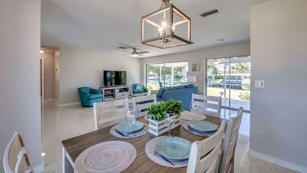 A dining area with a wooden table set for four, a ceiling fan, and a view of the outdoors through large windows. - Cozy in the Cape, Heated Saltwater Pool & Gulf access, water view  (Cape Coral)