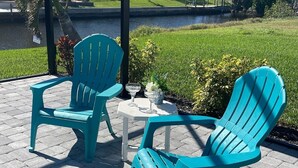 Two turquoise Adirondack chairs and a small round table with a glass on it, set on a paved patio with a grassy area and a water feature in the background. - Cozy in the Cape, Heated Saltwater Pool & Gulf access, water view  (Cape Coral)