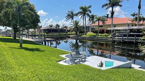 A well-maintained grassy area with a pool, lounge chairs, and a small deck overlooking a calm water body. - Cozy in the Cape, Heated Saltwater Pool & Gulf access, water view  (Cape Coral)