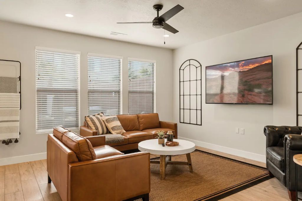 A living room with a brown leather sofa, a round coffee table, and a black leather armchair. There are three windows with blinds, a ceiling fan, and a large painting on the wall.