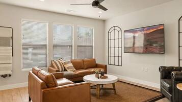 A living room with a brown leather sofa, a round coffee table, and a black leather armchair. There are three windows with blinds, a ceiling fan, and a large painting on the wall.