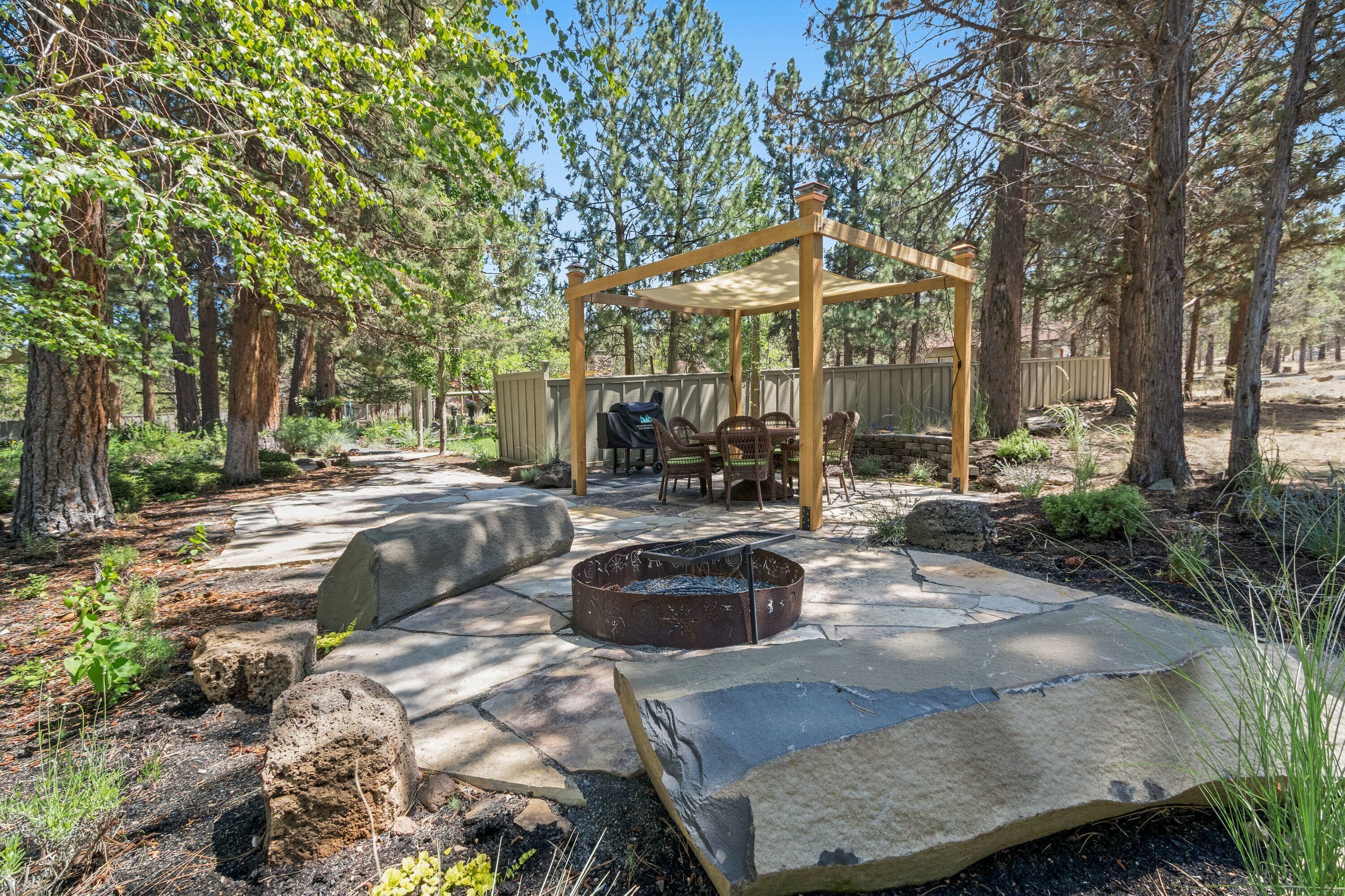 A covered outdoor seating area with a fire pit, surrounded by a stone patio and a wooden pergola.