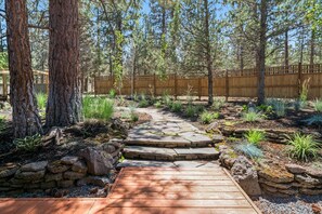 Une terrasse en bois qui mène à un sentier pavé de pierres, bordé d’arbres et de plantes.