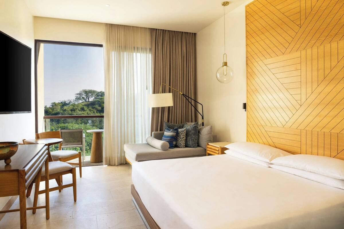 A hotel room with a bed, a desk with chairs, a sofa, a TV, and a view of greenery outside.