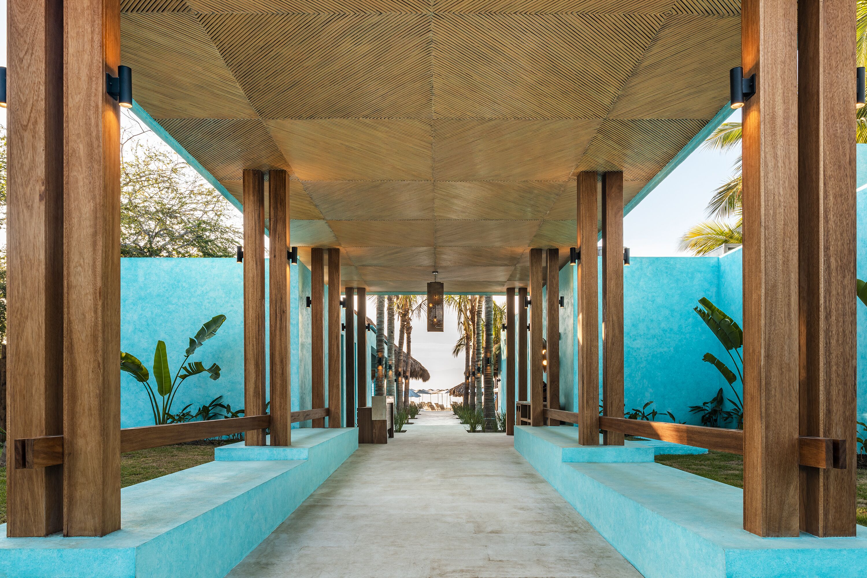 A covered walkway with wooden pillars and a blue wall, leading to a pool area.