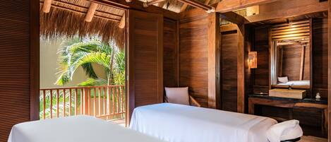 A room with two beds, a wooden ceiling, a thatched roof, a wooden bench, and a bathroom with a sink and mirror.