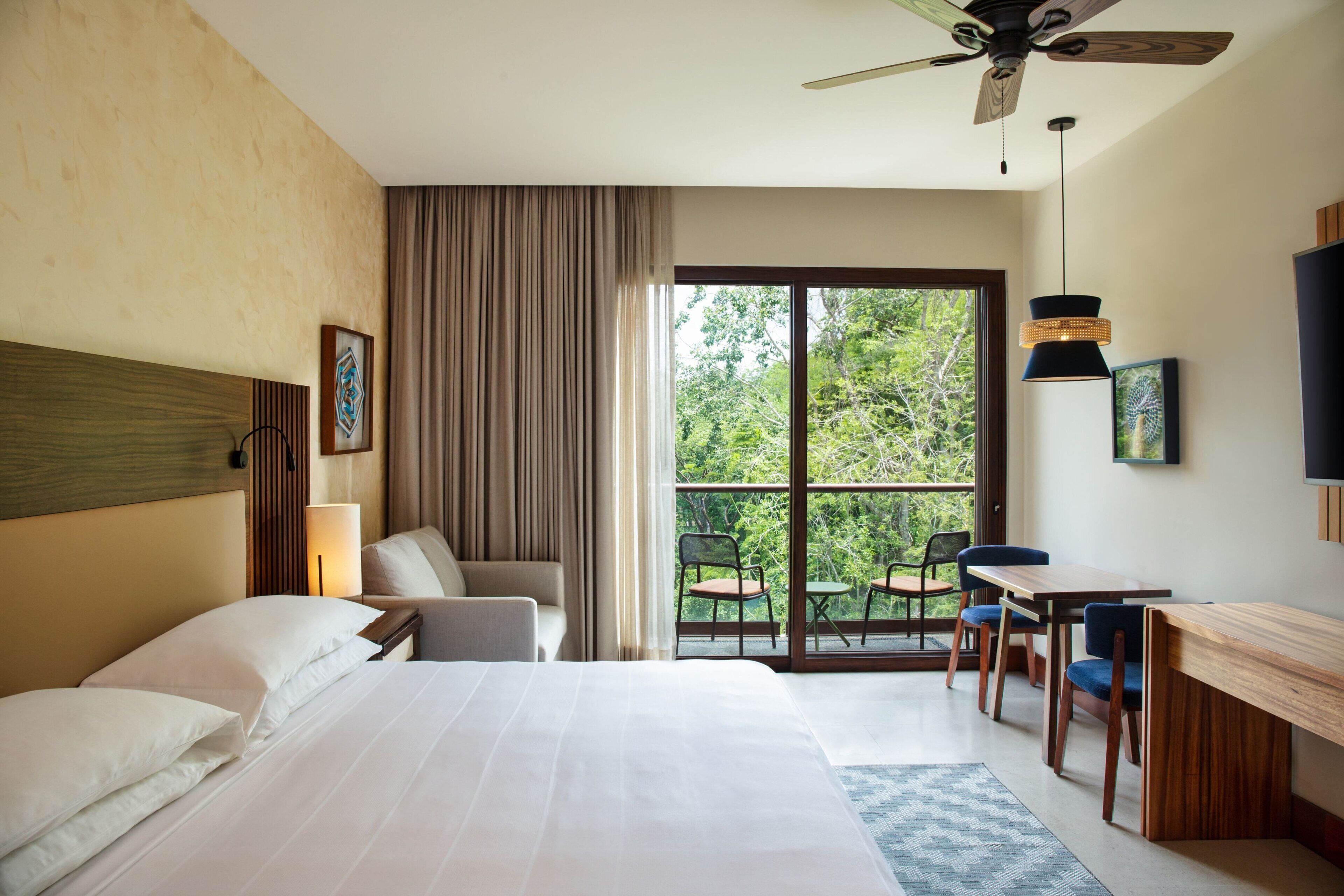 Quarto de hotel com cama, escrivaninha, cadeira, ventilador de teto e uma grande janela com vista para o verde.