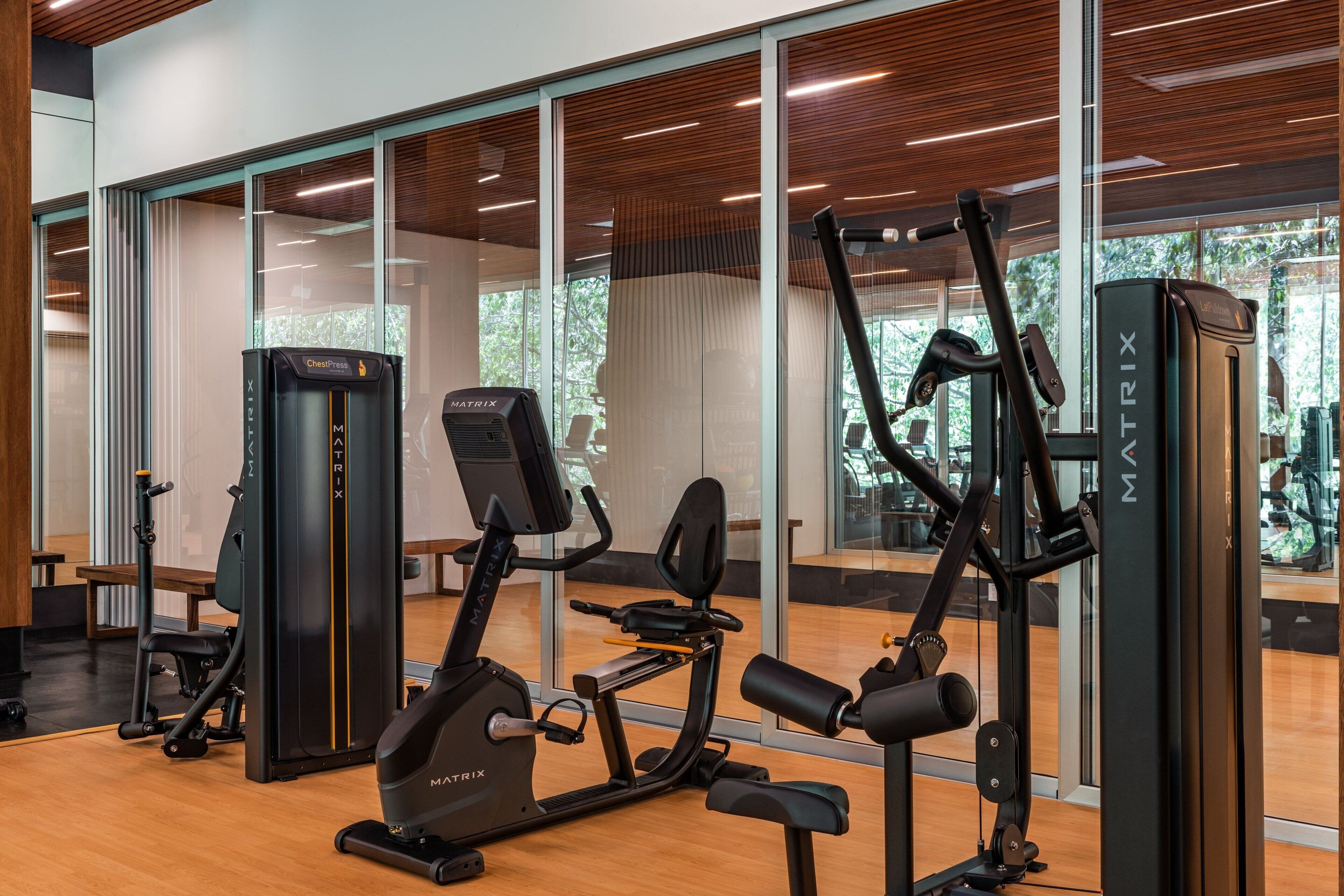 A gym with various exercise machines, large windows, and a wooden floor.