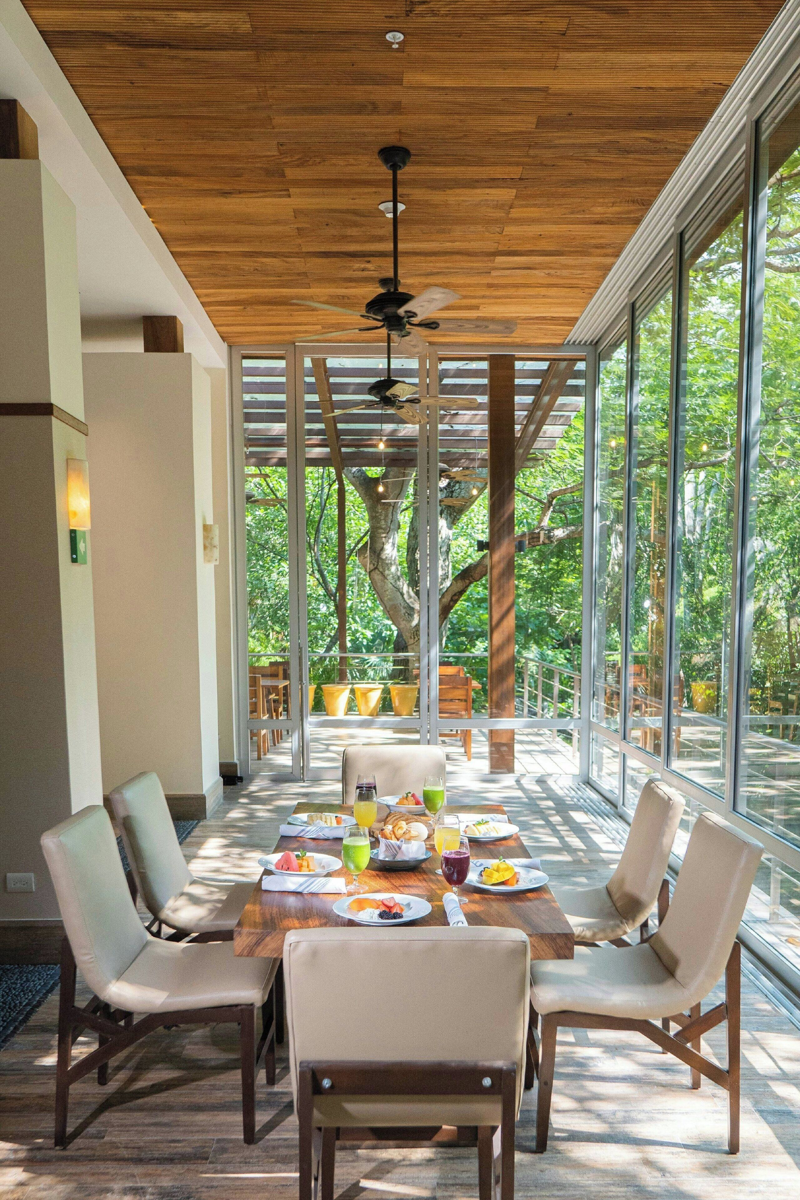 A dining area with a wooden ceiling, large windows, and a view of trees outside.