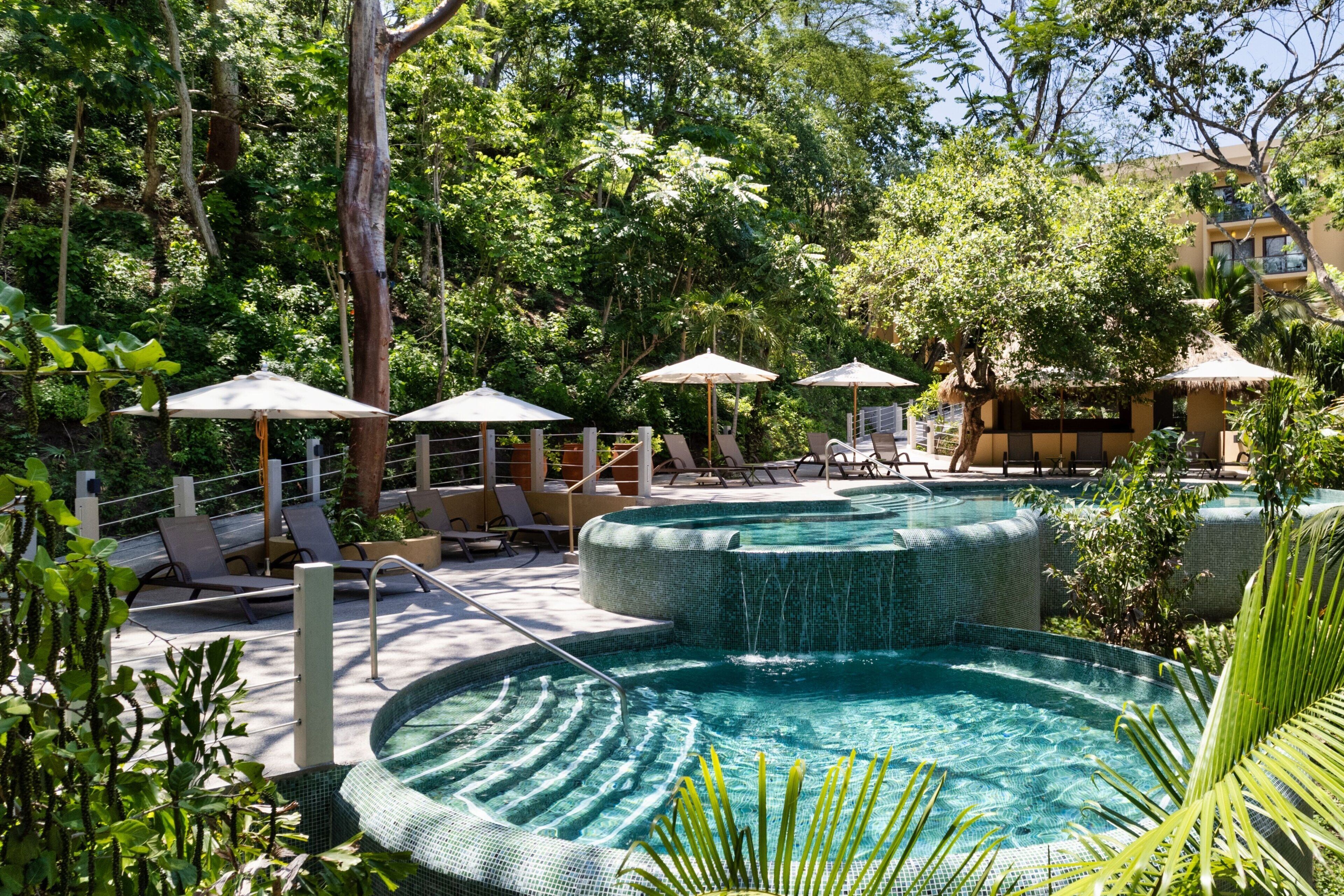 A pool area with lounge chairs and umbrellas, surrounded by lush greenery.
