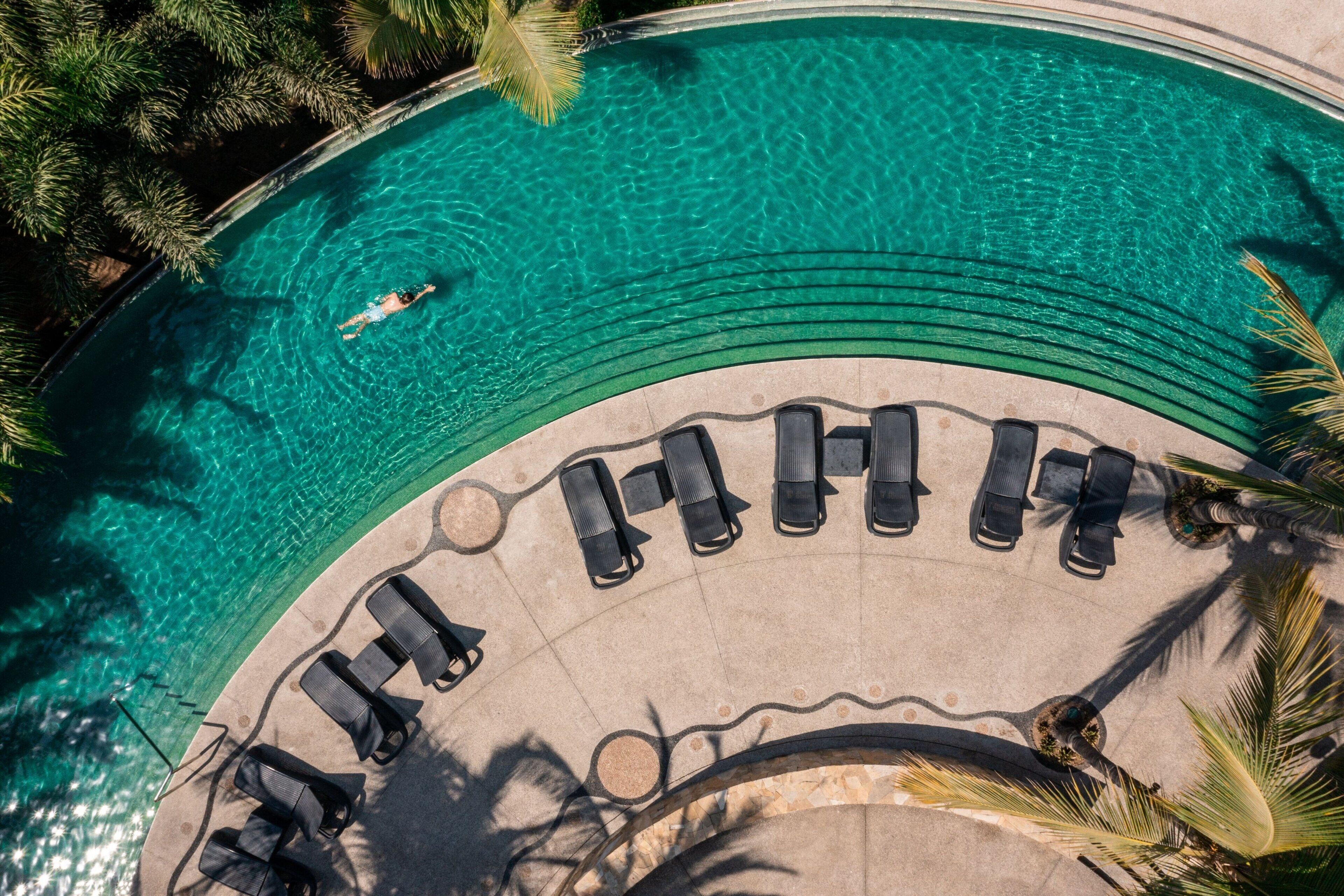 Vista aérea de una piscina con agua cristalina, rodeada de sillones de descanso y palmeras.