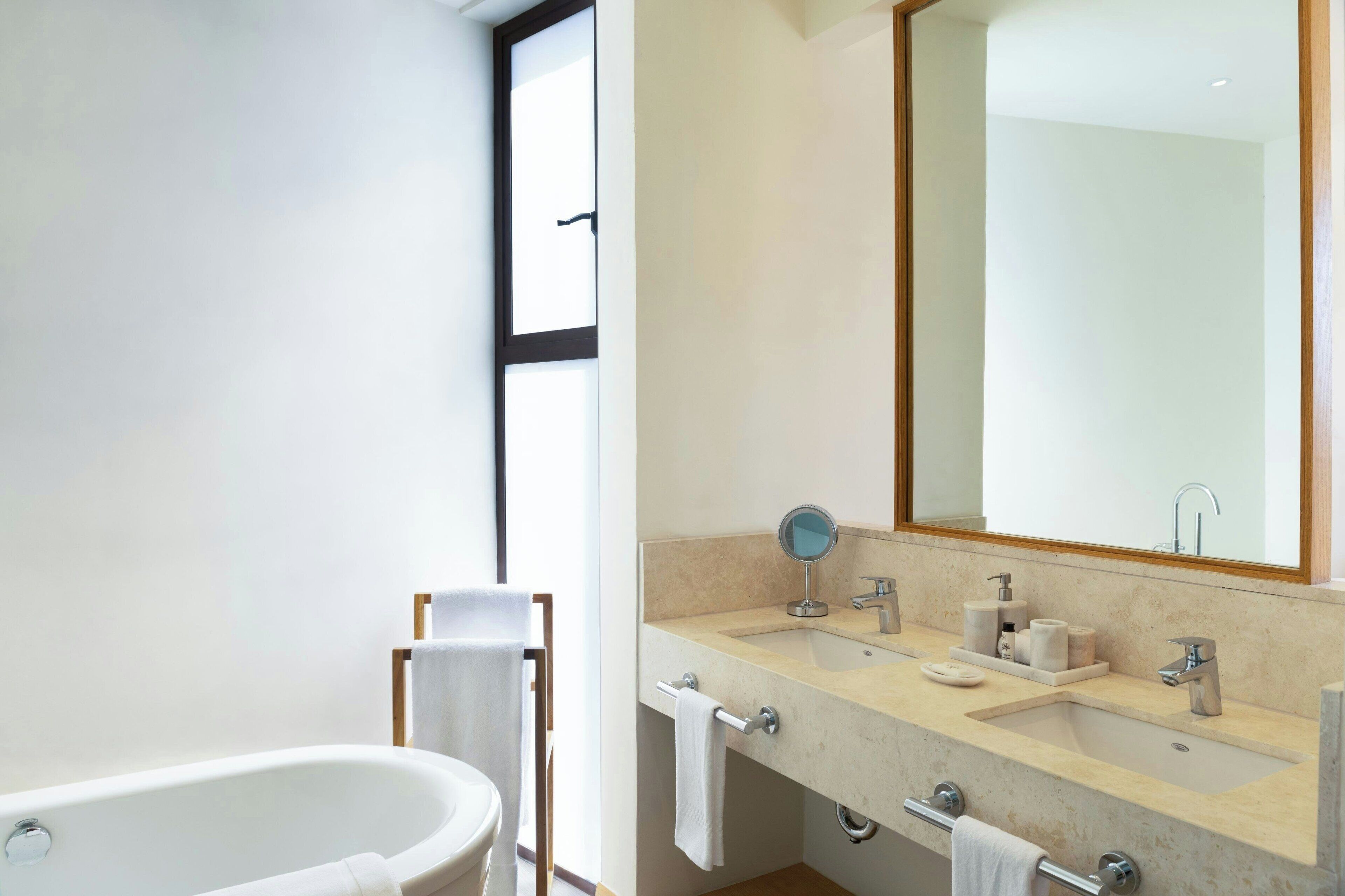A modern bathroom with a bathtub, two sinks, a large mirror, and a window.