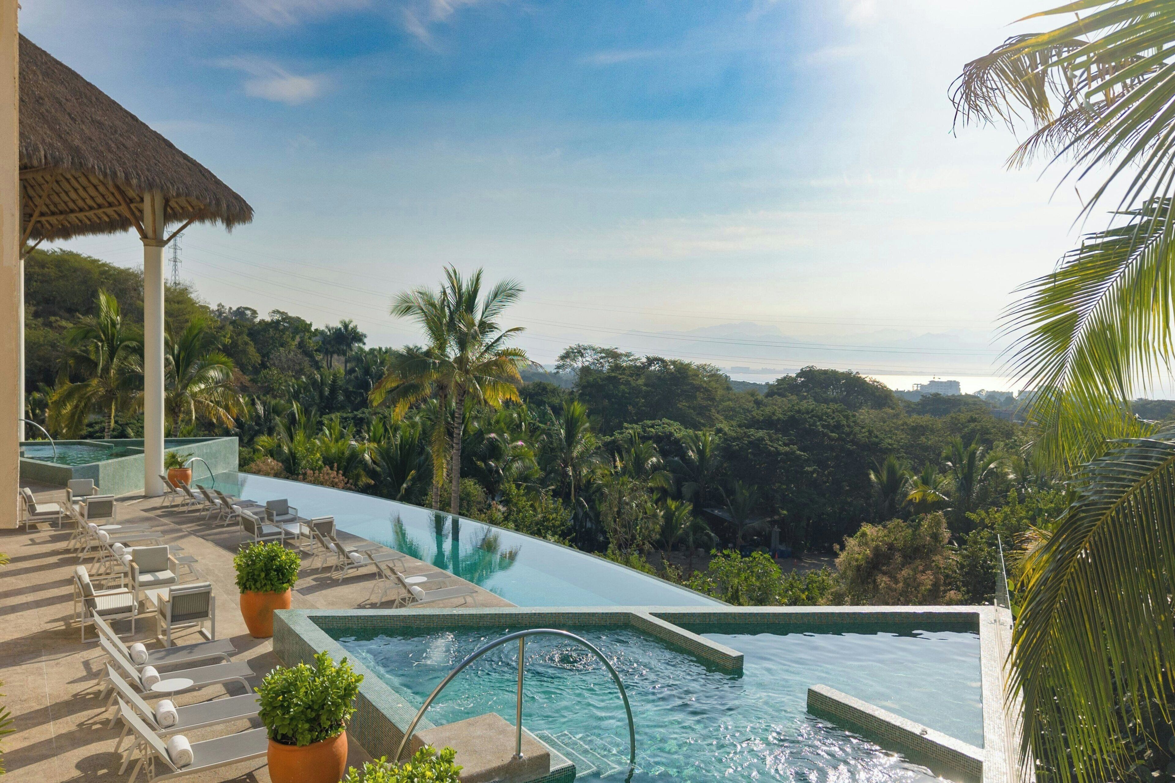Una piscina infinita con sillones de descanso y plantas en macetas, rodeada de exuberante follaje y vistas del horizonte de la ciudad.