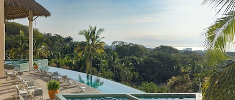 Una piscina infinita con sillones de descanso y plantas en macetas, rodeada de exuberante follaje y vistas del horizonte de la ciudad.