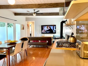 Eine moderne Küche mit Gasherd, Toaster und Wasserkocher. Es gibt eine Essecke mit Tisch und Stühlen sowie ein Wohnzimmer mit Sofa und Fernseher.