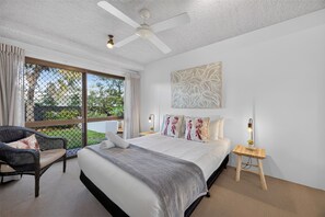 A bedroom with a bed, a chair, a nightstand, a ceiling fan, and a window with curtains. - Ground Floor Apt 800m to Hastings Street & Beach (Noosa Heads)