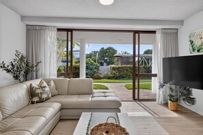 A modern living room with a sectional sofa, a flat-screen TV, and a large sliding glass door leading to a garden. - Ground Floor Apt 800m to Hastings Street & Beach (Noosa Heads)
