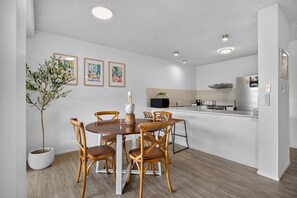 A modern kitchen with white cabinetry, a dining area with wooden chairs, and a potted plant. - Ground Floor Apt 800m to Hastings Street & Beach (Noosa Heads)