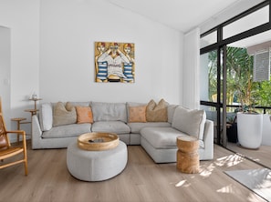 A modern living room with a sectional sofa, round ottoman, and a large window with a view of greenery. - The River View - 10 min walk to Noosa Beach (Noosa Heads)