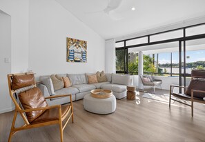 Ein modernes Wohnzimmer mit Ecksofa, Ledersessel, rundem Hocker und Seeblick durch große Fenster.