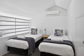 A modern, compact room with two beds, a window with blinds, an air conditioning unit, and a bedside table with a lamp.