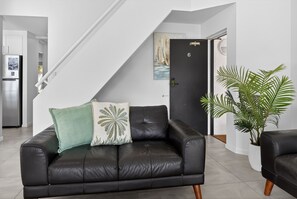 A modern living room with a black leather sofa, a green and white pillow, a potted plant, and a staircase leading to another room. - Premium 3Bed Apt - Walking distance to Hastings St (Noosa Heads)