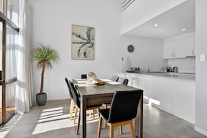 A modern kitchen with white cabinets, a dining area with a wooden table and black chairs, and a potted plant. - Premium 3Bed Apt - Walking distance to Hastings St (Noosa Heads)