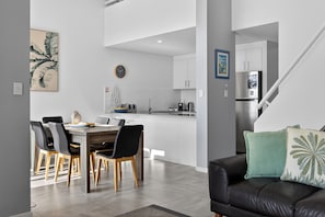 A modern kitchen with white cabinets, a stainless steel refrigerator, and a dining area with a wooden table and chairs. - Premium 3Bed Apt - Walking distance to Hastings St (Noosa Heads)