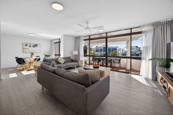 A modern living room with a sofa, dining table, and chairs, large windows with curtains, and a ceiling fan. - Noosa Parade Escape - 800m to Hastings Street! (Noosa Heads)