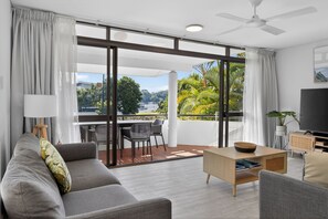 A modern living room with a sofa, coffee table, TV, and a balcony with outdoor furniture. - Noosa Parade Escape - 800m to Hastings Street! (Noosa Heads)